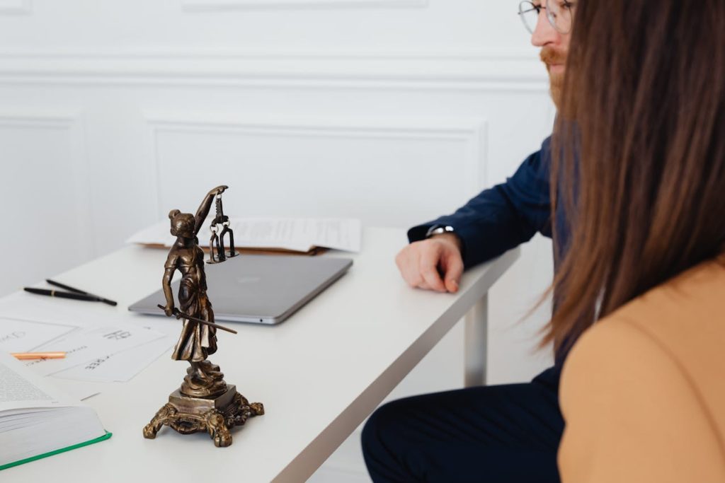 Estátua de bronze da Lady Justiça em uma mesa de escritório, cercada por laptop, papéis e canetas, em um ambiente profissional com duas pessoas ao fundo.