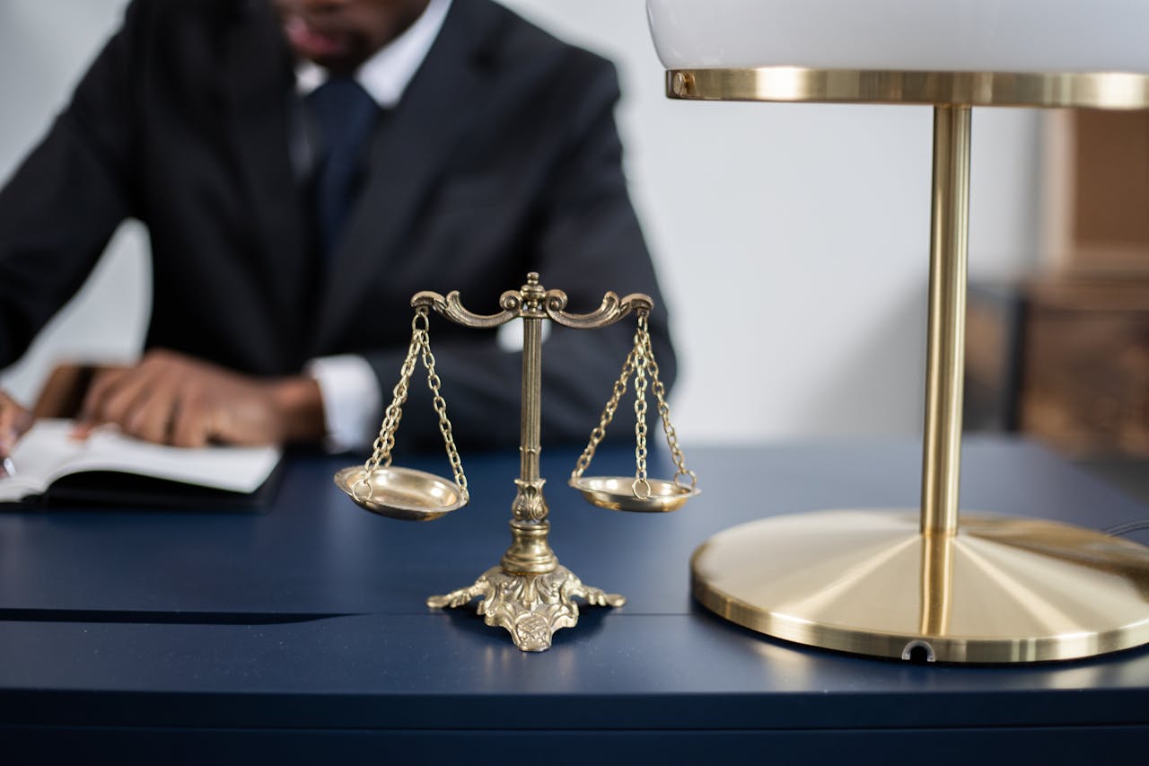 Escalas de justiça douradas em destaque sobre uma mesa de escritório, com uma pessoa ao fundo escrevendo em um documento, simbolizando um ambiente jurídico.